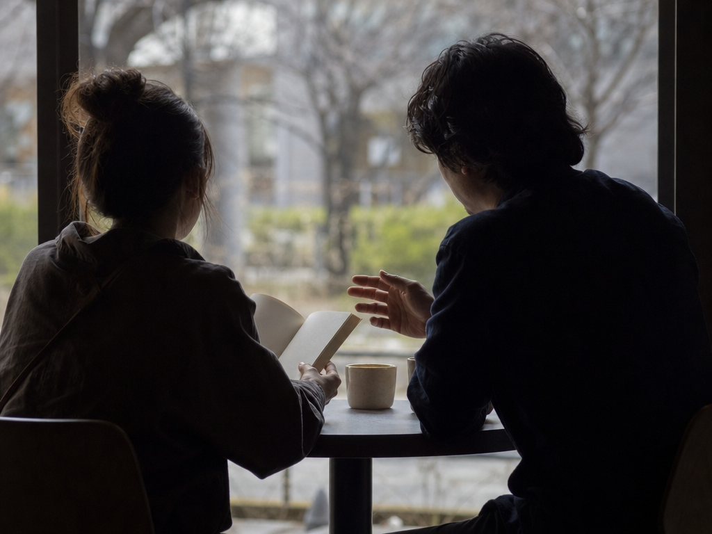 カフェの窓辺で資料を前に対話するふたりの人物の後ろ姿。テクニカル分析とファンダメンタル分析を比較検討する場面を象徴する情景